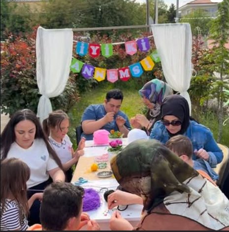Bahçede uzun bir masanın etrafında çocuklar, anneler ve öğretmenler birlikte el işi atölyesi yapıyor.